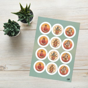 Op de afbeelding: Een vel met ronde stickers met kleurrijke illustraties van hindoe-goden. Elke sticker heeft een witte rand en een oranje en gele gradi&euml;nt achtergrond. Het vel ligt op een lichtgroene achtergrond, met twee kleine vetplanten in pot.
