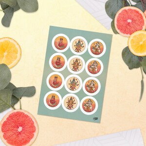 Op de afbeelding: Een vel stickers met ronde illustraties van goden. Elke sticker heeft een witte rand en een gedetailleerd, kleurrijk ontwerp met een goudgele achtergrond. De stickers zijn gerangschikt op een lichtblauwe achtergrond.