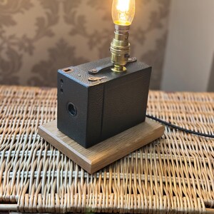 May include: A vintage camera converted into a table lamp with a light bulb and a wooden base. The lamp is sitting on a wicker surface.