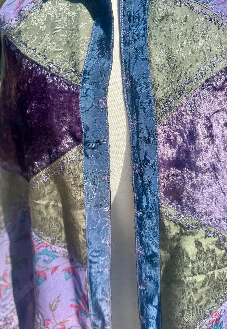 Pode incluir: Um casaco patchwork com tons de roxo, verde e azul. O casaco tem uma textura aveludada e padr&otilde;es florais. As bordas s&atilde;o aparadas com um tecido azul. O casaco &eacute; feito de v&aacute;rios tecidos.