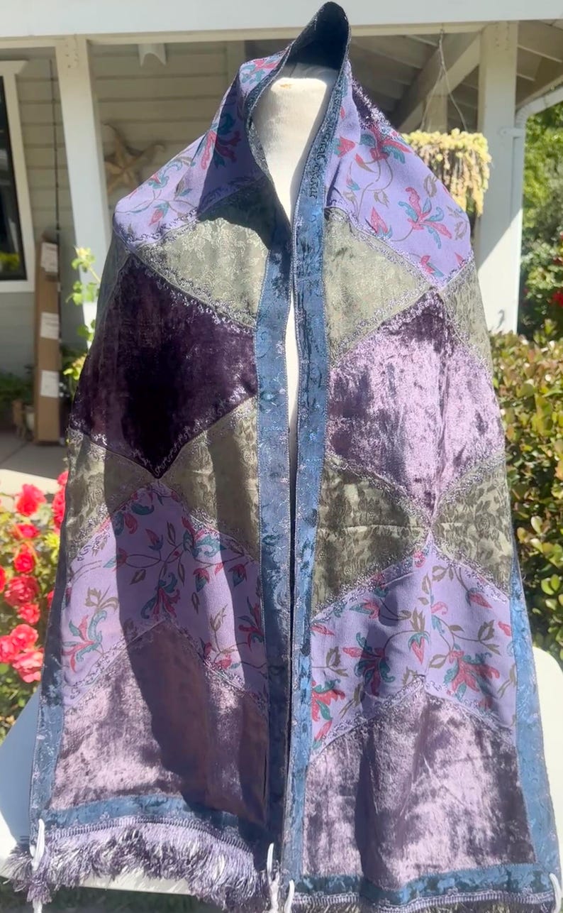 Pode incluir: Um xale de patchwork com uma variedade de tecidos em tons de roxo, verde e azul. O xale apresenta padr&otilde;es florais, veludo e uma borda com franjas. O design inclui um painel central e patches em forma de diamante.