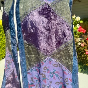Pode incluir: Um cachecol de patchwork com uma variedade de texturas e cores. O cachecol apresenta sec&ccedil;&otilde;es de veludo roxo, tecido verde-oliva e tecido lavanda estampado com desenhos florais. Acabamento azul e franjas completam o design.