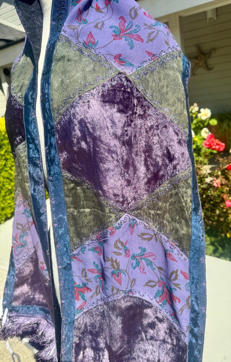 Pode incluir: Um cachecol de patchwork com uma variedade de texturas e cores. O cachecol apresenta sec&ccedil;&otilde;es de veludo roxo, tecido verde-oliva e tecido lavanda estampado com desenhos florais. Acabamento azul e franjas completam o design.