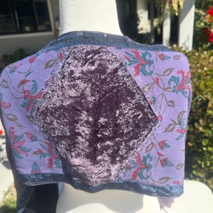 Pode incluir: Bolero roxo com um painel central de veludo e mangas estampadas com flores. As mangas s&atilde;o de um roxo mais claro com desenhos florais vermelhos, verdes e azuis. O casaco tem uma borda azul escura e texto ao longo da gola.