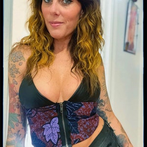 May include: A black and purple floral print corset top with a zipper closure. The top is worn over a black bra and paired with black jeans.