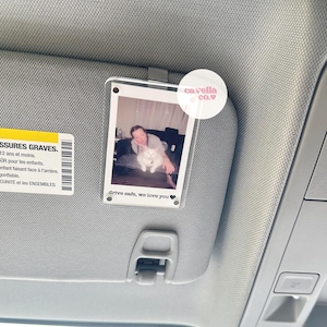 May include: A clear acrylic car visor clip with a Polaroid photo of a person holding a white cat. The photo is surrounded by a black border and has the text "drive safe, we love you" printed below it. A pink circle sticker with the text "cavella co." is attached to the top right corner of the clip.