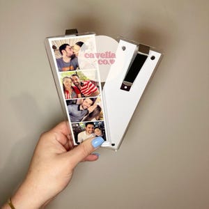 May include: A heart-shaped photo display with a clear plastic front and a white back. The display holds four vertical photo strips. The text "cavella co." is visible on the display. A black metal clip secures the display.