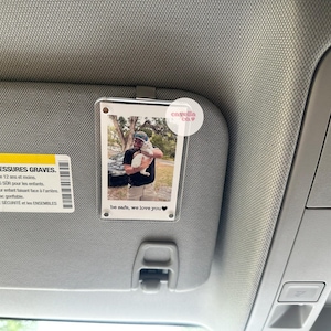 May include: A clear plastic photo holder with a white border, attached to a car's sun visor. The photo shows a man holding a white cat. The text "be safe, we love you" is printed on the photo holder.