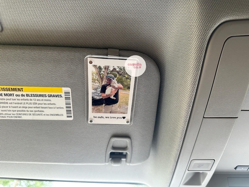 May include: A clear plastic photo holder with a white border, attached to a car's sun visor. The photo shows a man holding a white cat. The text "be safe, we love you" is printed on the photo holder.