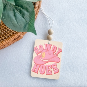 May include: A beige car air freshener with a pink cowboy hat and the text "Howdy Hoes".
