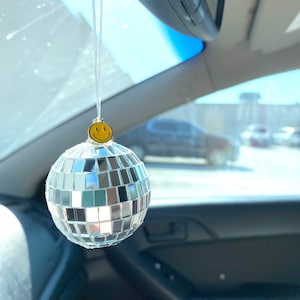 May include: A silver disco ball car charm with a yellow smiley face hanging from the rearview mirror of a car.