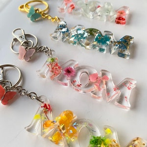 May include: Four keychains with the word "MOM" spelled out in clear resin with embedded dried flowers and glitter. Each keychain has a different color scheme and flower arrangement. The keychains are attached to silver key rings with butterfly charms.