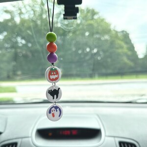 May include: A Halloween-themed car charm with a string of beads and three round charms. The charms feature a pumpkin, a black cat, and a ghost.