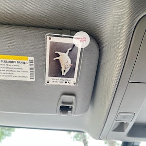 May include: A white cat with black paws is pictured in a clear plastic photo holder with a black clip. The photo holder is attached to a car's sun visor. The text "You left pawprints on my heart" is printed on the photo holder.