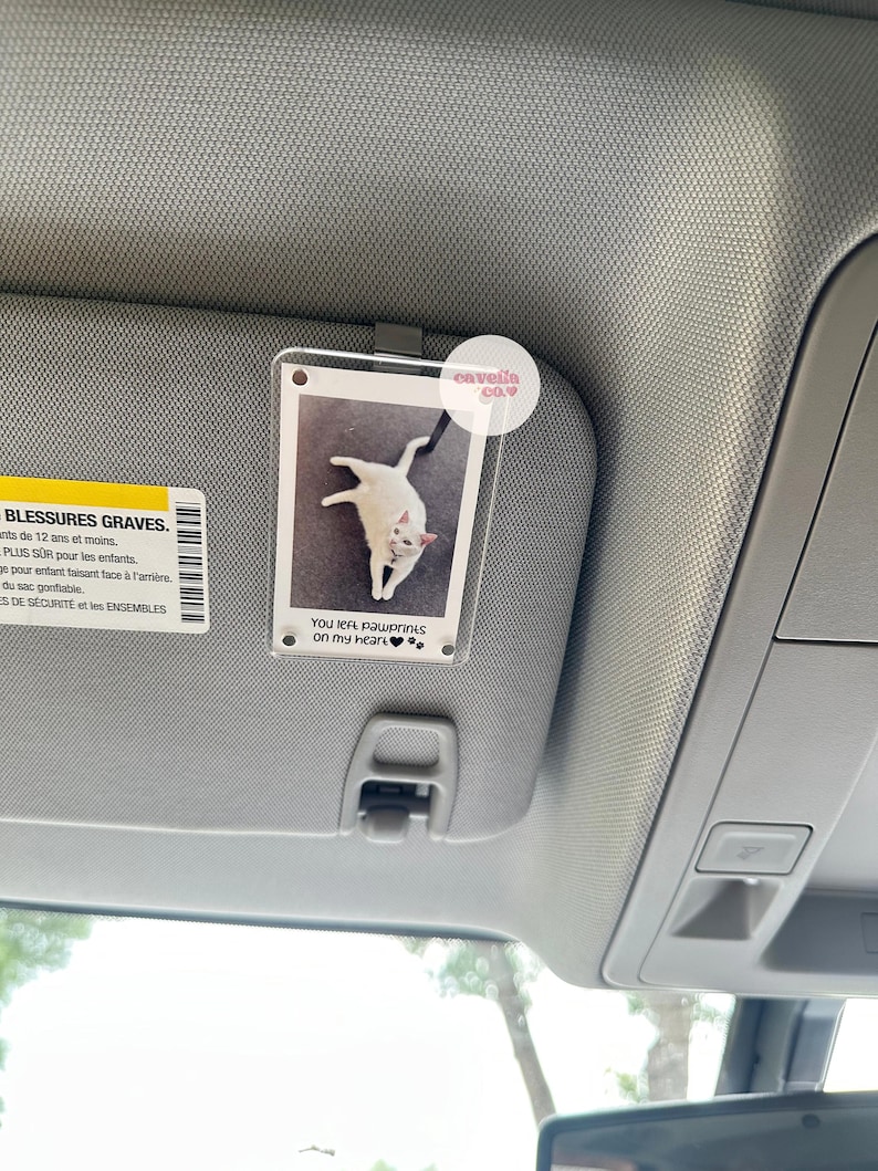 May include: A white cat with black paws is pictured in a clear plastic photo holder with a black clip. The photo holder is attached to a car's sun visor. The text "You left pawprints on my heart" is printed on the photo holder.