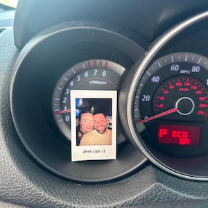 May include: A car's dashboard with a speedometer and tachometer. The speedometer reads 0 mph and the tachometer reads 0 rpm. A Polaroid photo of a couple is attached to the dashboard with the text "drive safe <3".