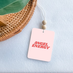May include: Pink car air freshener with white string and wooden beads. The air freshener has the text "ANGEL ENERGY" in red.