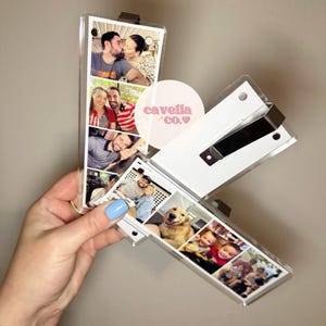 May include: A clear acrylic photo box with multiple family photos. The box is open, revealing the photos. The box has a white backing and a black metal clasp. The text "cavella co." is on the box.