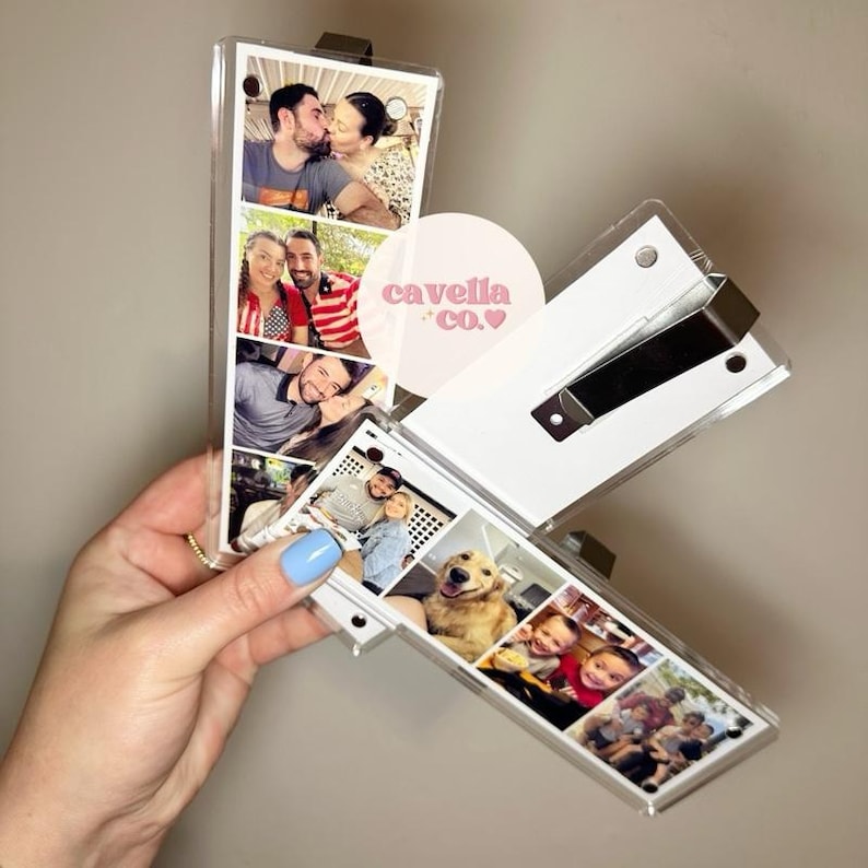 May include: A clear acrylic photo box with multiple family photos. The box is open, revealing the photos. The box has a white backing and a black metal clasp. The text "cavella co." is on the box.