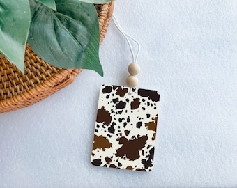May include: A white card with a brown and black cowhide pattern. The card is hanging from a white string with two wooden beads.