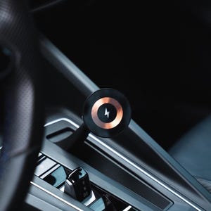 Può includere: Un supporto magnetico circolare per telefono da auto con bordo esterno nero e anello interno color rame metallizzato, caratterizzato da un'icona a forma di fulmine bianco al centro, posizionato su un braccio nero nella console centrale.
