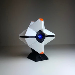 Puede incluir: Un objeto geométrico blanco y negro con detalles en naranja y una luz azul en el centro. El objeto está sobre una base negra. El diseño recuerda una estética futurista de ciencia ficción.