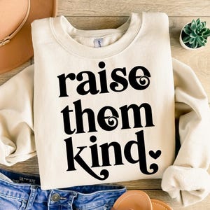Peut inclure: Sweat-shirt crème avec l'inscription "raise them kind" en grandes lettres noires stylisées. Le sweat-shirt est plié sur une surface en bois, avec un jean bleu et un chapeau visibles en arrière-plan. Une petite plante succulente est également présente.