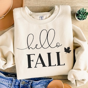 May include: Cream-colored sweatshirt with the words "hello FALL" in black script and block letters, along with a black maple leaf graphic. The sweatshirt is folded on a wooden surface, with a hat and jeans visible.