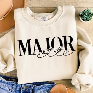 May include: Cream-colored sweatshirt with the words "MAJOR elite" in black script. The sweatshirt is folded, with a pair of blue jeans and a brown hat visible in the background. A small succulent plant is in the upper right corner.