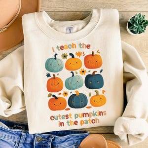 Puede incluir: Sudadera de color crema con un gráfico colorido de nueve calabazas sonrientes en varios tonos de naranja, azul y amarillo. El texto "I teach the cutest pumpkins in the patch" está impreso debajo. La sudadera se combina con unos vaqueros azules.