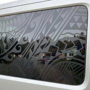 May include: A rectangular vehicle window featuring a dark grey adhesive decal with a complex pattern of geometric shapes, triangles, and curved lines. The design covers the glass surface, creating a semi-transparent effect over the view.
