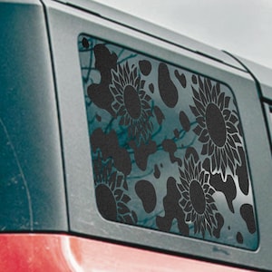 May include: Black sunflower window decal applied to a vehicle window. The decal features several large sunflowers and abstract shapes. The vehicle is dark gray and red.