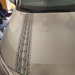 May include: A silver car hood featuring a vertical black decal with a repeating geometric tribal pattern. The decal runs from the base of the windshield toward the front of the vehicle, displaying sharp, angular lines and symmetrical shapes.