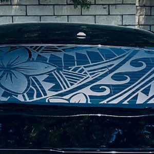May include: A black car rear window decal featuring a large white plumeria flower on the left side, integrated into a complex Polynesian-inspired tribal pattern with geometric shapes, triangles, and wave motifs covering the entire glass surface.