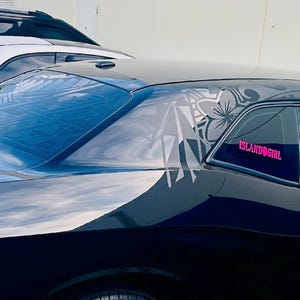 May include: A black coupe car featuring a large grey floral decal on the rear side window and roof pillar. A bright pink decal with the text ISLANDGIRL is applied to the side window glass. The vehicle has a sleek, dark exterior finish.
