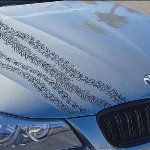 May include: Silver car hood with black floral design. The hood features a BMW emblem and a black grill. The floral design is composed of interconnected leaves and flowers, creating a decorative pattern across the hood.