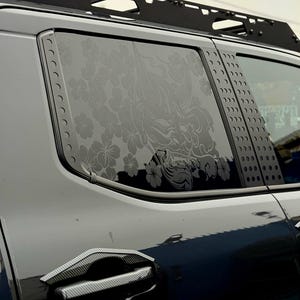 May include: A dark gray vehicle window decal featuring a detailed Hannya mask surrounded by cherry blossom flowers. The decal is applied to the rear side window of a vehicle, which also features black metal pillar trim and a roof rack system.