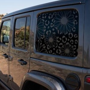 May include: A dark gray SUV features a rear side window decorated with a black decal. The decal displays a repeating pattern of large daisy flowers layered over a leopard print background, covering the entire surface of the glass window.