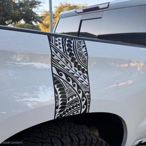 May include: A white pickup truck features a vertical black vinyl decal applied to the rear quarter panel. The decal displays a detailed Polynesian-inspired tribal pattern consisting of geometric shapes, triangles, and wave-like motifs.