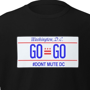 Puede incluir: Camiseta negra con un gráfico rectangular. El gráfico tiene el texto "Washington, D.C." encima de las palabras "GO = GO" en azul. Debajo del texto está "#DONT MUTE DC" en blanco.