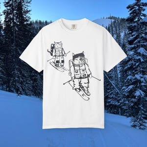 Puede incluir: Camiseta blanca con una ilustración en blanco y negro de dos gatos esquiando. Los gatos llevan equipo de esquí y mochilas. El texto "SNOW CAT" es visible en la chaqueta de uno de los gatos. El fondo muestra una escena de montaña nevada.