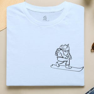 Puede incluir: Camiseta blanca con una ilustración en blanco y negro de un gato practicando snowboard. La camiseta tiene cuello redondo y una pequeña ilustración de gato cerca del cuello. La ilustración del gato se repite en la parte delantera.