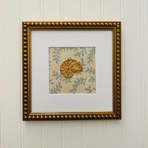 May include: A framed artwork featuring a gold nautilus shell on a blue and white floral patterned fabric background.