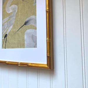 May include: Framed artwork featuring a watercolor painting of white herons on a textured, golden-yellow background. The artwork is encased in a gold bamboo-style frame, adding a touch of elegance. The frame is mounted on a white, vertical-paneled wall.