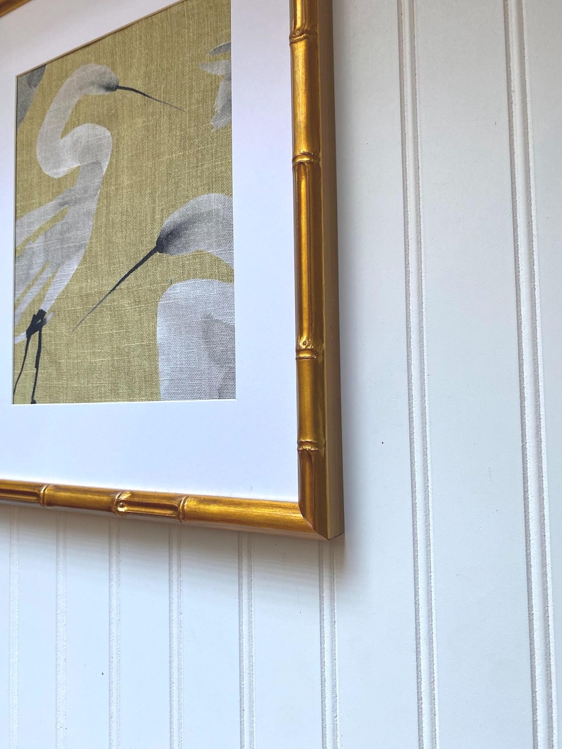 May include: Framed artwork featuring a watercolor painting of white herons on a textured, golden-yellow background. The artwork is encased in a gold bamboo-style frame, adding a touch of elegance. The frame is mounted on a white, vertical-paneled wall.