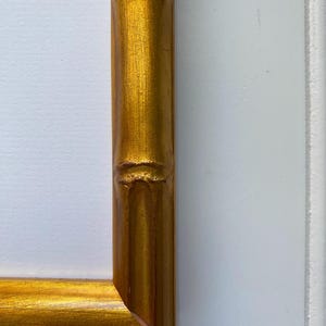 May include: Close-up of a gold-colored picture frame corner. The frame has a bamboo-like design with a textured surface. The frame is against a white background.
