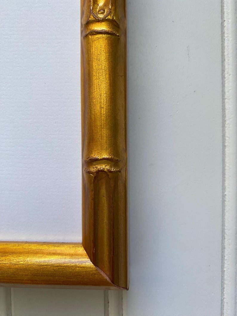 May include: Close-up of a gold-colored picture frame corner. The frame has a bamboo-like design with a textured surface. The frame is against a white background.