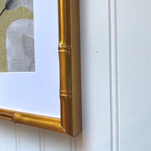 May include: A framed artwork featuring a gold bamboo-style frame. The artwork within the frame depicts a white and gray floral design against a muted yellow-green background. The frame is a warm gold color, adding a touch of elegance.