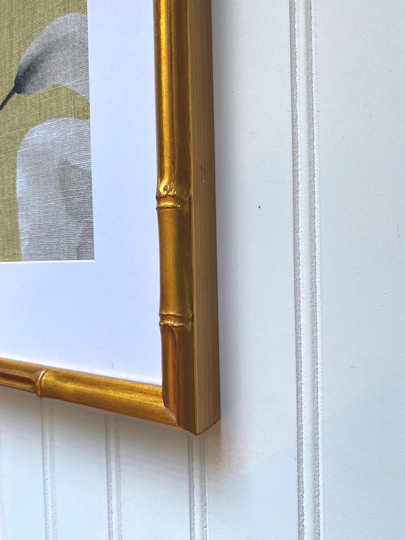May include: A framed artwork featuring a gold bamboo-style frame. The artwork within the frame depicts a white and gray floral design against a muted yellow-green background. The frame is a warm gold color, adding a touch of elegance.