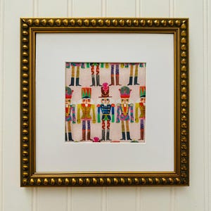 May include: A framed print of a colorful pattern featuring nutcracker soldiers in various poses. The print is set against a light pink background and is framed in a gold frame with a beaded border.